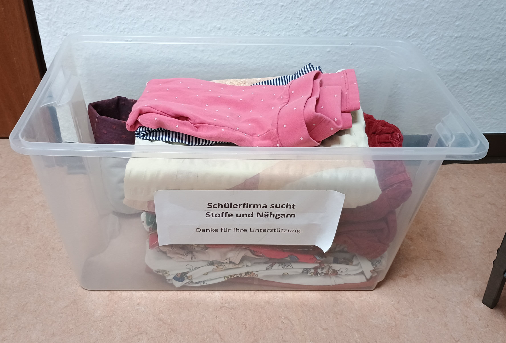 Bild einer transparenten Sammelbox. Darin sind Stoffspenden (T-Shirts, Pullis, Nähgarn, etc...)